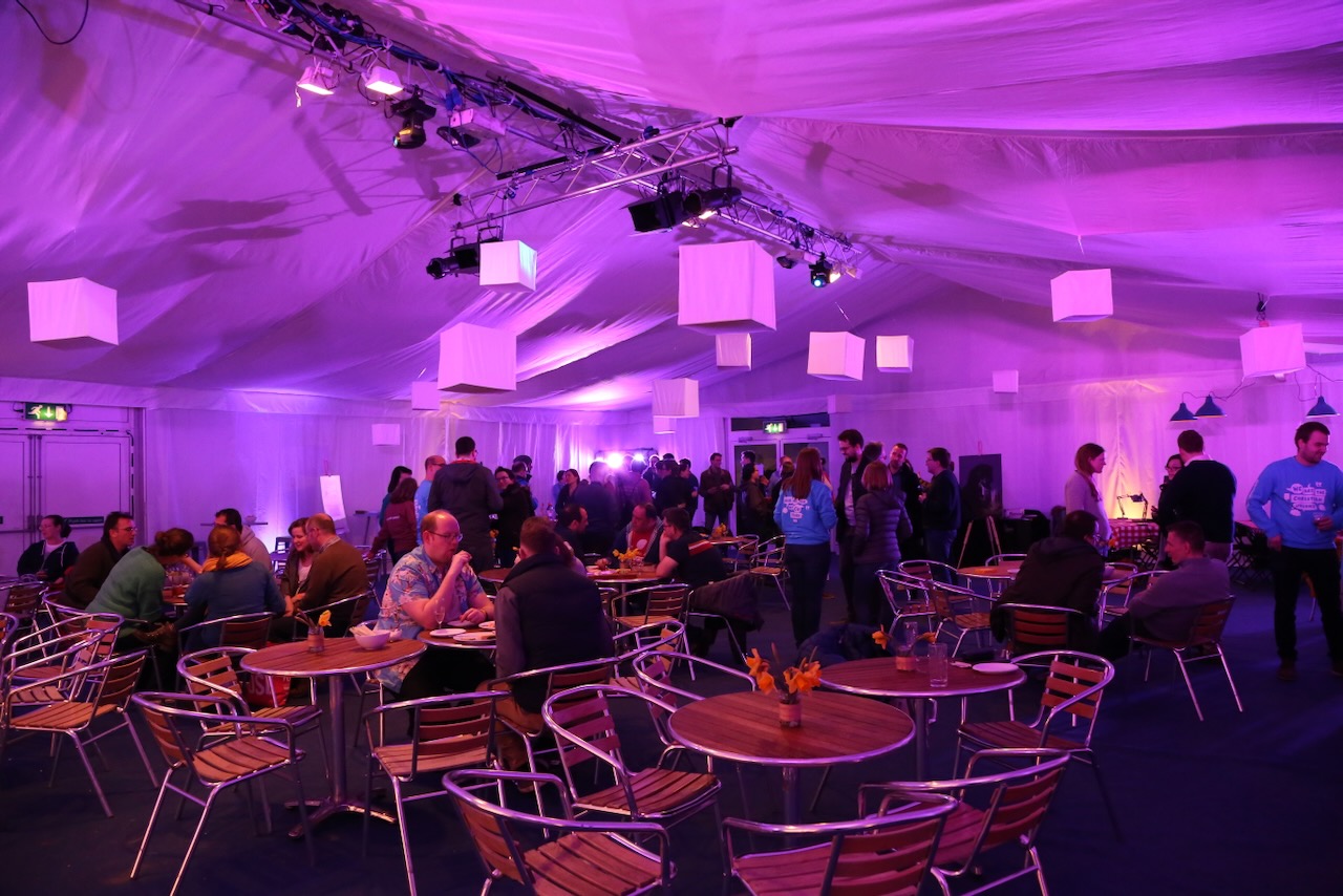 A social event with people stood and sat at tables with mauve lighting
